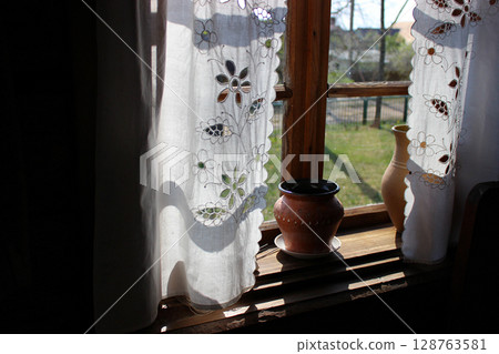 Sunlight filtering through delicate curtains at a rustic window with potted plants in a tranquil outdoor setting during late afternoon hours 128763581