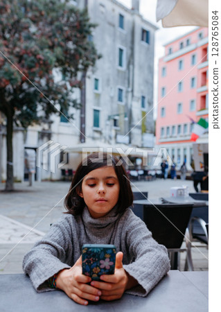 a child sitting at the table outdoors and watching the phone 128765084