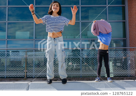 girl in striped t-shirt and jeans jumping next to a smaller girl who is taking off sweatshirt 128765122