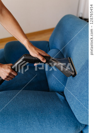 Vertical shot of cleaning technician operating industrial vacuum cleaner, sanitizing upholstered blue furniture within contemporary residential interior, maintaining spotless living space, close-up. Vertical shot of cleaning technician operating industrial vacuum cleaner, sanitizing upholstered blue furniture within contemporary residential interior, maintaining spotless living space, close-up. 128765454