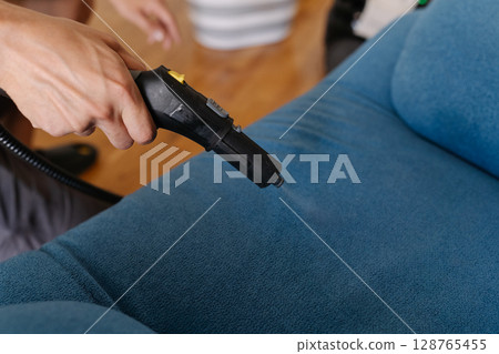 Housewife sanitizing blue upholstered sofa using powerful steam cleaner, targeting deep dirt removal and eliminating household bacteria, close-up. Concept of domestic work, housekeeping and household. Housewife sanitizing blue upholstered sofa using powerful steam cleaner, targeting deep dirt removal and eliminating household bacteria, close-up. Concept of domestic work, housekeeping and household. 128765455