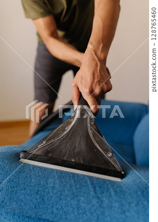 Vertical cropped shot of unrecognizable housewife male using wet vacuum cleaner to remove dirt and stains from blue sofa, ensuring hygienic cleaning process. Concept of cleanliness and housekeeping. 128765460