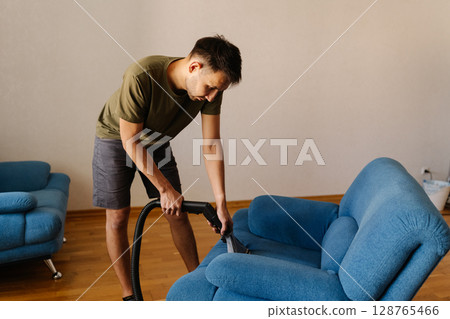 Portrait of cleaning service worker male in casual clothes using washing vacuum cleaner to clean blue sofa in modern living room with wooden floor. Concept of cleanliness and housekeeping. Portrait of cleaning service worker male in casual clothes using washing vacuum cleaner to clean blue sofa in modern living room with wooden floor. Concept of cleanliness and housekeeping. 128765466