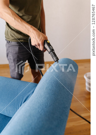 Vertical cropped shot of cleaning service employee sanitizing blue sofa with high-temperature steam cleaner in contemporary living space, removing dirt and bacteria. Concept of hygiene and cleanliness Vertical cropped shot of cleaning service employee sanitizing blue sofa with high-temperature steam cleaner in contemporary living space, removing dirt and bacteria. Concept of hygiene and cleanliness 128765467