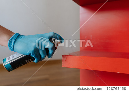 Housewife wearing blue gloves applying scotch remover spray to dried adhesive stain on red shelf, performing thorough housekeeping at home, close-up. Concept of easy housekeeping domestic life. 128765468