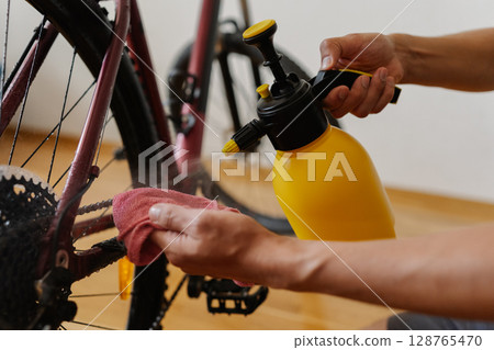 Detail cropped shot of unrecognizable man using yellow sprayer and cloth preparing to clean chain of mountain bike, emphasizing importance of bicycle maintenance after trail riding at home. 128765470