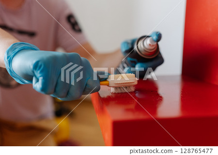 Cropped shot of housewife male wearing blue protective gloves removing adhesive stains, scrubbing red wooden shelf with small brush during detailed cleaning process at home, close-up. Cropped shot of housewife male wearing blue protective gloves removing adhesive stains, scrubbing red wooden shelf with small brush during detailed cleaning process at home, close-up. 128765477