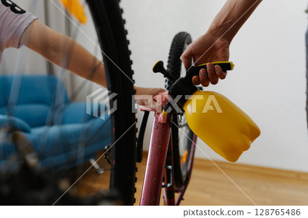 Two MTB biker male cleaning mountain bike using water sprayer and cloth, removing dirt and grime from frame at workshop, performing detailed maintenance after trail riding, close-up. 128765486