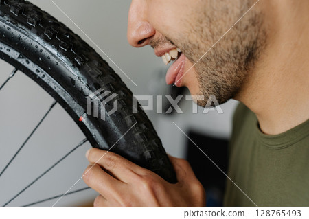 Close-up cropped shot cheerful man licking tire of mountain bike wheel while holding rag, showcasing humorous approach to bicycle maintenance, side view portrait. Concept of bicycle transport service. 128765493