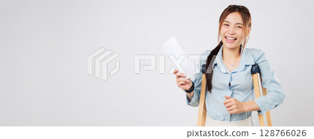 Injured Asian woman leg or foot broken holding bill payment paper and standing on crutches, smiling while using crutches for support after accident and recovery. isolated studio white background 128766026