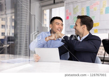 Teammates giving fist bump gesture seated at desk with tablet Teammates giving fist bump gesture seated at desk with tablet 128767001