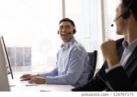 Asian man, callcenter employee laughing seated at desk with colleague 128767173
