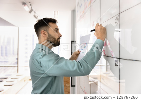 Serious man holding papers with charts and writing on whiteboard 128767260