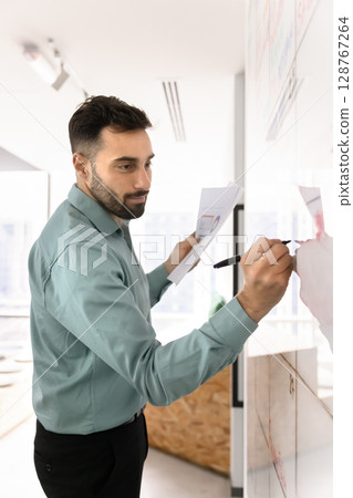 Hispanic man holds papers, writing information on whiteboard Hispanic man holds papers, writing information on whiteboard 128767264