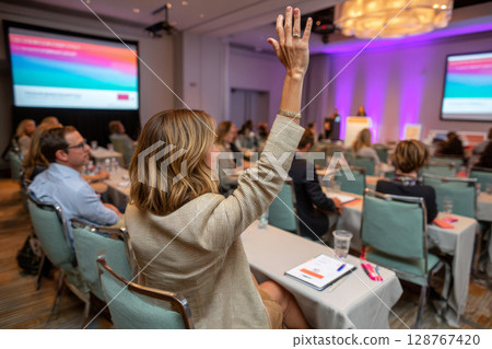 Businesswoman raising hand in modern conference room during corporate presentation with presenter and slides Businesswoman raising hand in modern conference room during corporate presentation with presenter and slides 128767420