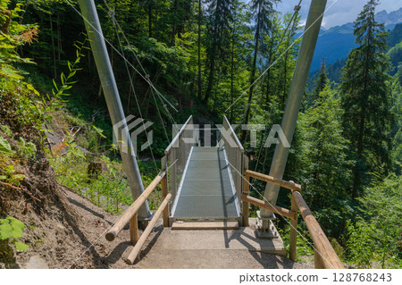 Steel suspension bridge on a forest trail near Partnachklamm, Garmisch-Partenkirchen, Bavaria. A modern walkway offering stunning views of pristine alpine nature and lush green woodland. 128768243