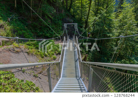 Steel suspension bridge on a forest trail near Partnachklamm, Garmisch-Partenkirchen, Bavaria. A modern walkway offering stunning views of pristine alpine nature and lush green woodland. 128768244