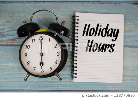 Holiday Hours text written on notepad with alarm clock on wooden background 128770656