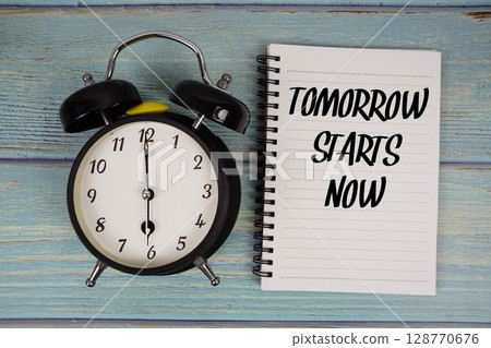 Tomorrow Starts Now text written on notepad with alarm clock on wooden background Tomorrow Starts Now text written on notepad with alarm clock on wooden background 128770676