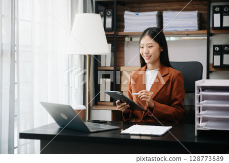Young beautiful woman typing on tablet and laptop while sitting at the working wooden table modern office 128773889