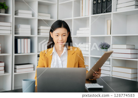 Confident Asian woman with a smile standing holding notepad and tablet at the office. 128774583