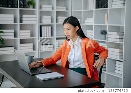 Beautiful Asian woman using laptop and tablet while sitting at her working place. Concentrated at work. Beautiful Asian woman using laptop and tablet while sitting at her working place. Concentrated at work. 128774713