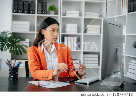 Beautiful Asian woman using laptop and tablet while sitting at her working place. Concentrated at work. 128774718