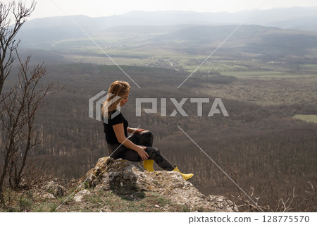 Woman Hillside Landscape: Blonde woman sits on a rock, viewing the vast valley landscape, daytime. Woman Hillside Landscape: Blonde woman sits on a rock, viewing the vast valley landscape, daytime. 128775570