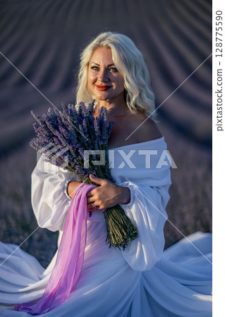 Blonde woman poses in lavender field at sunset. Happy woman in white dress holds lavender bouquet. Aromatherapy concept, lavender oil, photo session in lavender 128775590