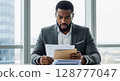 A concerned businessman reviews important documents, his expression reflecting worry and surprise at his desk near a large window. 128777047