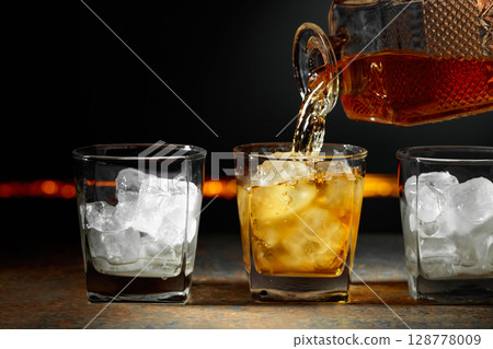 Whiskey is poured from a decanter into a glass with ice. 128778009
