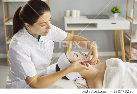 Young attractive cosmetologist woman applying sheet mask on female face in salon 128778283