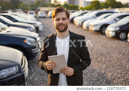A smiling car salesman in a suit holding a tablet in a car dealership, with rows of cars in the background. A smiling car salesman in a suit holding a tablet in a car dealership, with rows of cars in the background. 128778372