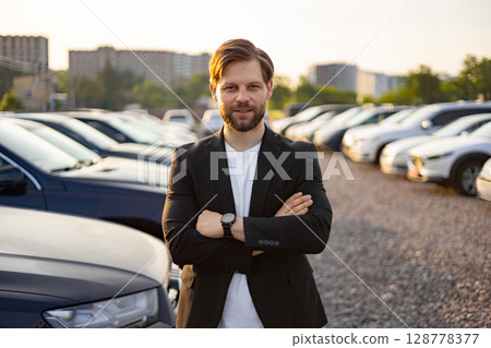 A confident businessman stands arms crossed at a car dealership, showcasing the available vehicles. The sun sets in the background. 128778377