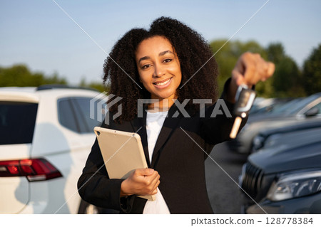 A smiling woman in a car dealership is selling a car, holding the keys and a tablet. A smiling woman in a car dealership is selling a car, holding the keys and a tablet. 128778384