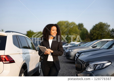 An African American woman in a car dealership holding a tablet, surrounded by vehicles. She is smiling, likely helping a customer. An African American woman in a car dealership holding a tablet, surrounded by vehicles. She is smiling, likely helping a customer. 128778387