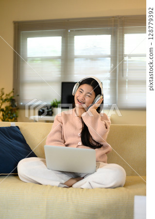Smiling teenage girl wearing headphones while enjoying music and using her laptop at home 128779392