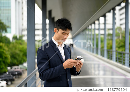 Businessman reading messages on mobile phone during break in city walkway 128779504