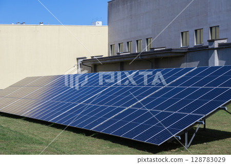 Expansive blue solar panel array on green grass by modern industrial buildings, efficiently generating clean renewable energy for sustainable enterprise power needs Expansive blue solar panel array on green grass by modern industrial buildings, efficiently generating clean renewable energy for sustainable enterprise power needs 128783029