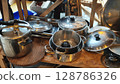 Collection of assorted metallic cooking pots and pans displayed on a rustic wooden shelf at a flea market in a lively urban setting 128786326