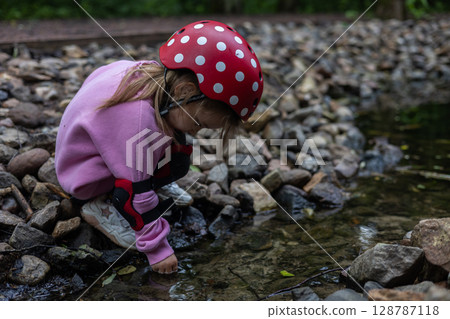 A curious child in protective gear, including a red helmet with white dots and elbow pads, explores nature by touching the cool water of a shallow stream. A child explores a small rocky stream. 128787118