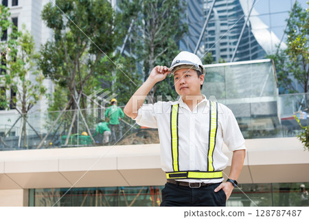 Lifting his white safety helmet slightly, an Asian foreman in a reflective vest looks ahead confidently at a construction site. Lifting his white safety helmet slightly, an Asian foreman in a reflective vest looks ahead confidently at a construction site. 128787487