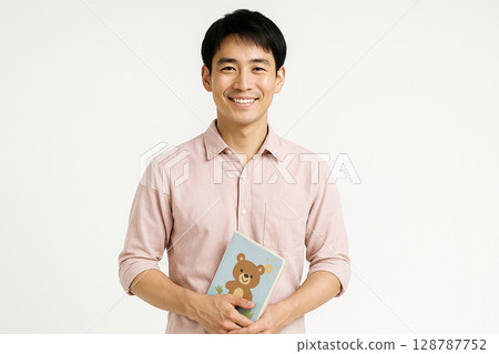 Smiling portrait of a young Japanese male nursery teacher holding a picture book White background 128787752