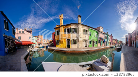Brightly colored houses reflect in the serene canal of Burano, Venice during a clear sunny day 128788963