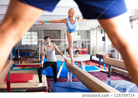 Woman gymnast performs complex acrobatic exercise on pommel horse with support of trainer in gym 128789714