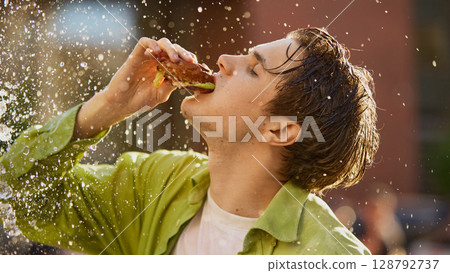 Wet-haired man in green shirt dramatically eating burger under splash Wet-haired man in green shirt dramatically eating burger under splash 128792737