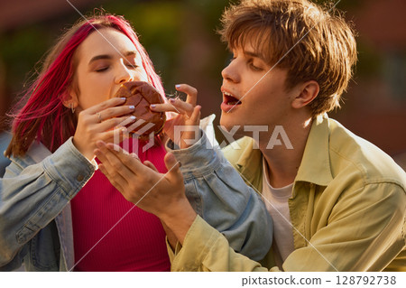 Young woman taking huge bite of burger while young man reacts with surprise Young woman taking huge bite of burger while young man reacts with surprise 128792738