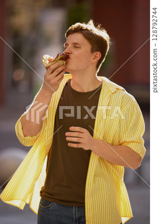 Young man taking a bite from a burger with passion in sunny street setting 128792744