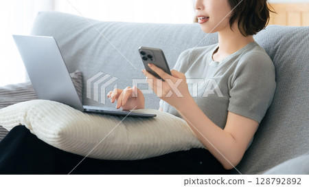 A woman sitting on a sofa and using a smartphone and a computer 128792892