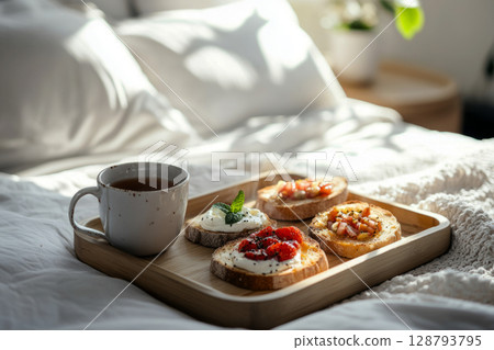 A delightful breakfast board features toasted bread topped with creamy spread and herbs, accompanied by a cup of hot tea, set against soft bed linens and natural light 128793795